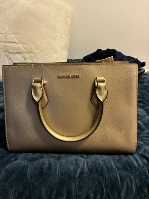 Guldig handväska från Michael Kors - Stilren handväska i guldigt skinn från Michael Kors. Väskan har två rejäla handtag, diskret logga i guld framtill och en strukturerad, fyrkantig form. Perfekt för dig som vill ha en elegant accessoar med plats för det viktigaste.