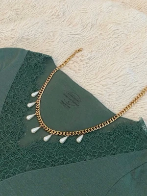Guldhalsband med droppformade pärlor - Snyggt halsband i guldfärgad metall med kedja och fem droppformade vita pärlor som hänger längs kedjan. Perfekt för att ge en elegant touch till din outfit. Passar till många olika stilar och är lätt att matcha med andra accessoarer.