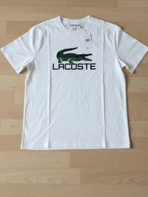 Vit Lacoste t-shirt med logga - Snygg vit t-shirt från Lacoste med den klassiska gröna krokodilloggan och texten 'LACOSTE' på bröstet. T-shirten har rund hals och korta ärmar. Perfekt till jeans eller shorts för en clean och stilren look.