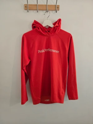 Orange hoodie från Peak Performance - Snygg orange hoodie från Peak Performance med vit logga på bröstet och liten logga på huvan. Hoodien har långa ärmar, huva och är tillverkad i ett mjukt material som känns skönt mot huden. Perfekt för en avslappnad och sportig stil.