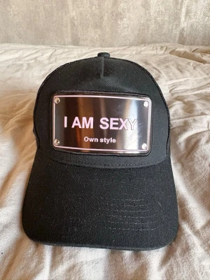 Svart keps med metallskylt - Säljer en svart keps med böjd skärm och mesh på sidorna. Framtill sitter en metallskylt med texten 'I AM SEXY Own style' i vitt. Kepsen har justerbar snapback och cool streetwear-vibe. Perfekt för dig som vill sticka ut.