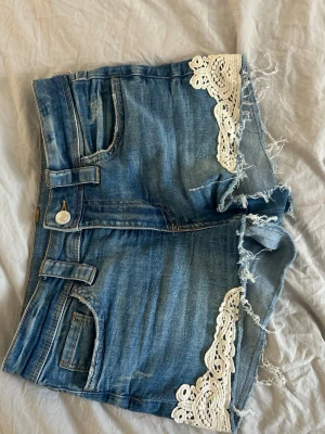 Blå jeansshorts med spetsdetaljer - Säljer ett par blå jeansshorts med råa kanter och vita spetsdetaljer vid bensluten. Shortsen har klassisk femficksdesign, knappgylf och en snygg, avslappnad look. Perfekta för varma dagar och ger en cool touch till din outfit.