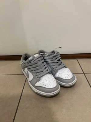 Nike Dunk Low gråvita sneakers - Nike Dunk Low sneakers i vitt och ljusgrått läder. Klassisk siluett med perforerad tåbox, grå snörning och vit sula. Swoosh-logga på sidan och vadderad häl för extra komfort. Perfekta för dig som gillar stilrena och tidlösa sneakers.