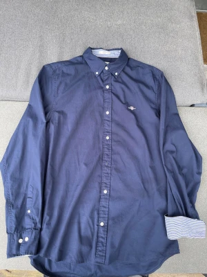 Mörkblå skjorta från Gant - Klassisk mörkblå skjorta från Gant med button-down krage och vita knappar. Skjortan har lång ärm och ett broderat Gant-emblem på bröstet. Insidan av kragen och manschetterna har randigt tyg för en snygg kontrast. Perfekt till jeans eller chinos.
