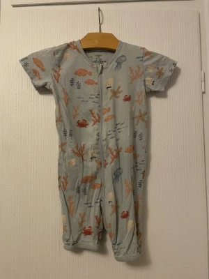 Blå kortärmad pyjamas Lindex, havsmotiv - Supersöt blå kortärmad pyjamas från Lindex med dragkedja framtill. Tryck med havsmotiv som fiskar, maneter, krabbor och sjöstjärnor i mjuka färger. Tillverkad i 90% bomull och 5% elastan för extra komfort. Perfekt för mysiga kvällar!