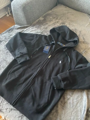  Ralph Lauren kofta  - Snygg svart hoodie från Polo Ralph Lauren med dragkedja framtill och klassisk logga broderad på bröstet. Hoodien har huva med dragsko, ribbade muddar och fickor på sidorna. Tillverkad i mjukt bomullsmaterial för en skön känsla. Storlek S och fraktar den under 24h