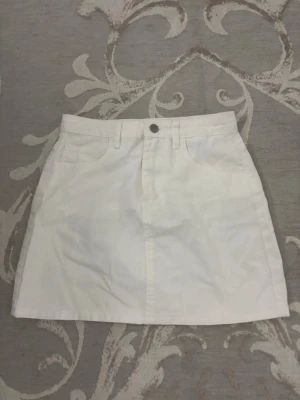 Vit jeans kortkjol  - Stilren vit kortkjol i jeans tyg med klassisk femficksdesign och knapp framtill. 
