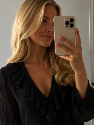 Svart blus med volang från Zara - Snygg svart blus från Zara med djup V-ringning och bred volangkant längs halsen. Blusen har långärmade ärmar med lätt puff och är gjord i ett tunt, skirt material som ger en luftig känsla. Perfekt för att lyfta vilken outfit som helst.