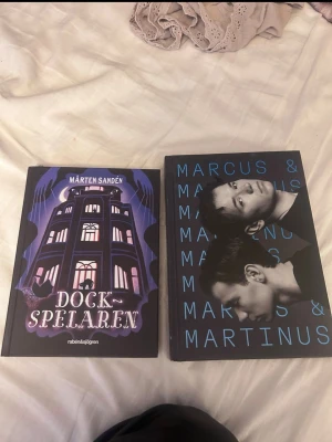 Dockspelaren & Marcus & Martinus bok - Två böcker: 'Dockspelaren' av Mårten Sandén med ett mystiskt, illustrerat omslag i lila och svart, samt en bok om Marcus & Martinus med svart omslag och blå text samt porträtt av tvillingarna. Perfekt för dig som gillar spänning och musik.