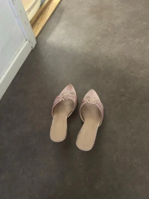 Rosa slip-in pumps med smal klack - Säljer ett par ljusrosa slip-in pumps med spetsig tå och smal klack. Skorna har dekorativa guldfärgade detaljer framtill och en vadderad rem över foten. Materialet ser ut att vara konstläder eller satinliknande tyg. Perfekta för dig som gillar en chic och trendig look.
