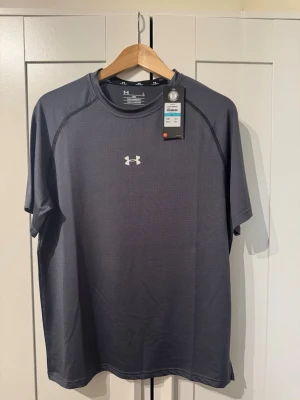 Mörkgrå tränings-tshirt Under Armour - Snygg mörkgrå tränings-tshirt från Under Armour med kort ärm och rund hals. Tillverkad i ett lätt och snabbtorkande funktionsmaterial, perfekt för träning. Diskret logga på bröstet och nacken. Skön passform den är i storlek L men kan passa M 
