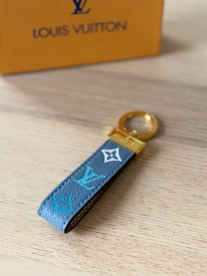  Vuitton nyckelring blå monogram - Säljer en snygg Vuitton nyckelring i blått läder med klassiskt monogrammönster i turkos och vitt. Nyckelringen har guldfärgad metall och exklusiv känsla, perfekt att hänga på väskan eller nycklarna.