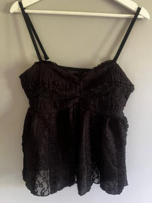 Shein Svart spetslinnetopp med tunna band - Säljer en svart linnetopp med tunna axelband och transparent spetsdetalj. Toppen har en luftig passform och vacker struktur i tyget, perfekt för dig som gillar en feminin och trendig stil.