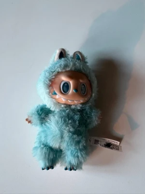 Pop Mart blå fluffig figur - Söt samlarfigur från Pop Mart med ett blått fluffigt monsterdräkt och stora blå ögon. Ansiktet är i plast och kroppen är täckt av mjukt, lurvigt tyg. Dräkten har små öron och tassar, och figuren har en etikett kvar.