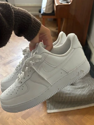 Vita Nike Air Force 1 sneakers - Klassiska helvita Nike Air Force 1 sneakers med låg profil. Skorna har en chunky sula, perforerad tå och ikonisk Swoosh-logga på sidan. Tillverkade i läder med vita skosnören och diskreta detaljer. Perfekta för en clean och stilren look.
