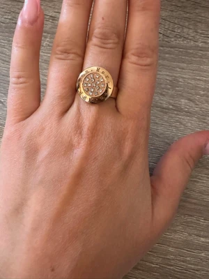 Guldfärgad ring med stenar från LenMer - Snygg guldfärgad ring med rund form och glittrande stenar på ovansidan. Ringen har en bred kant med ingraverad text runt om och en platt, dekorerad topp. Perfekt accessoar för att lyfta din stil. Ringen är i storlek 7 och helt oanvänd! OBS: FRAKTAR INOM 24H!🩷