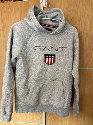 Grå hoodie från GANT med logga - Snygg gråmelerad hoodie från GANT med broderad logga och sköld på bröstet. Klassisk känguruficka framtill och huva. Tillverkad i mjukt bomullsmaterial och har ribbade muddar vid ärmslut och nederkant. Perfekt för en avslappnad och sportig stil.