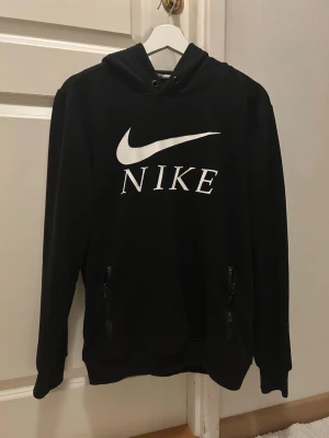 Svart hoodie med Nike-logga - Svart hoodie från Nike med stor vit logga och text på bröstet. Tröjan har huva med dragsko och två fickor med dragkedja framtill. Klassisk passform och ribbade muddar vid ärmar och nederkant. Perfekt för en sportig och avslappnad stil. Storlek M