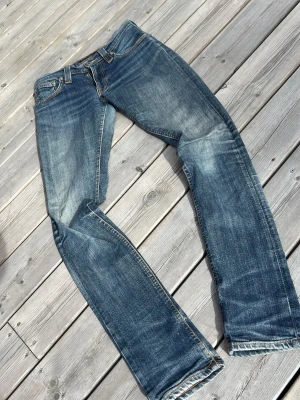 Blå jeans från Nudie Jeans Co - Säljer ett par klassiska blå jeans från Nudie Jeans Co med snygg slitning och raka ben. Jeansen har fem fickor, orangea sömmar och är gjorda i denim. Perfekta för en avslappnad och trendig stil.