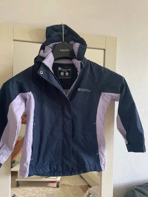  vindjacka från Mountain warehouse  - Snygg vindjacka i mörkblått och ljuslila med huva och dragkedja. Jackan har sportig design med kontrastpaneler och justerbara ärmslut. Perfekt för blåsiga dagar och enkel att matcha med olika outfits.