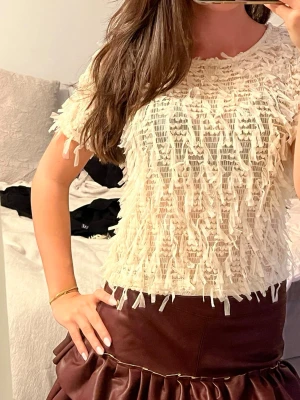 Krämvit topp med fransar - Säljer en krämvit topp med kort ärm och coola fransdetaljer över hela plagget. T-shirten har rund halsringning och är tillverkad i ett lätt tyg som ger en unik struktur. Perfekt för dig som vill sticka ut med en trendig och lekfull stil.