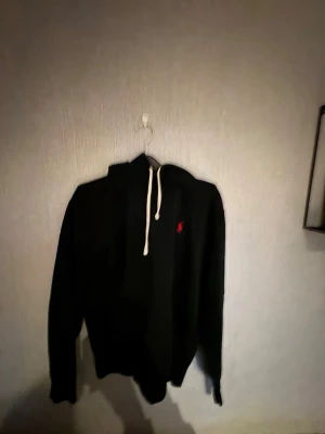 Svart hoodie från Polo Ralph Lauren - Svart hoodie från Polo Ralph Lauren med vit dragsko i huvan och den klassiska röda loggan broderad på bröstet. Hoodien har en avslappnad passform och är tillverkad i mjukt material som passar perfekt till en chill stil. 