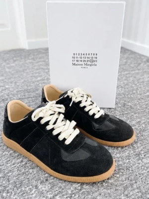 Maison Margiela svarta sneakers - Maison Margiela sneakers i svart mocka och läder med vita snören och beige gummisula. Klassisk low-top modell med stilren design och diskreta detaljer. Perfekta för dig som gillar minimalistisk och trendig stil.