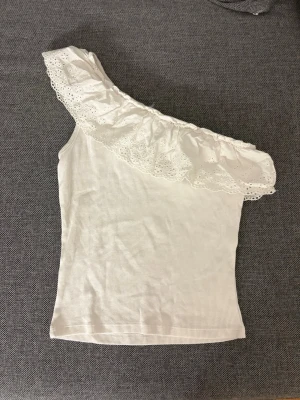 Vit oneshoulder topp med volang - Superfin vit oneshoulder topp med bred volang och broderad spetsdetalj längs axeln. Ribbad struktur i materialet och perfekt för en trendig och somrig look. Sitter tight och ärmlös med en axel bar. Inte säker på storleken!!!💕