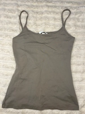 Mörk grå  linnetopp från H&M - Säljer en enkel och stilren beige linnetopp från H&M. Toppen har tunna axelband och är figurnära i modellen, perfekt att matcha med jeans eller kjol. Materialet är mjukt och skönt bomull som känns bekvämt mot huden.