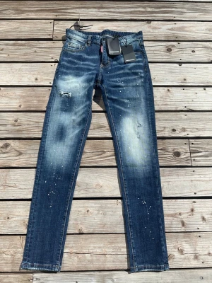 Blå slim fit jeans Dsquared2 - Säljer ett par blå slim fit jeans från Dsquared2 med slitningar, ljusa tvättade partier och färgstänk för en cool, trendig look. Klassisk femficksmodell med låg midja och smala ben. Perfekta för dig som gillar streetwear och vill sticka ut.