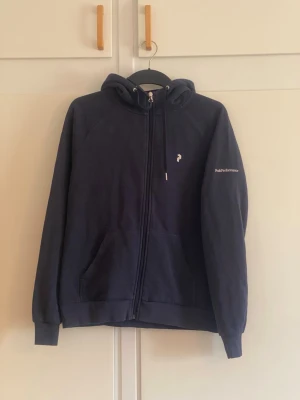 Mörkblå hoodie från Peak Performance - Mörkblå hoodie med dragkedja från Peak Performance. Klassisk modell med huva och dragsko, två fickor framtill och logga på bröstet samt ärmen. Perfekt för en avslappnad och sportig stil.