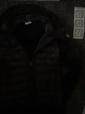 Svart dunväst från Moncler - Snygg svart dunväst från Moncler med klassisk quiltad design och dragkedja framtill. Västen har Monclers ikoniska logotyp på bröstet och en hög krage som ger extra stilpoäng. Perfekt för lager-på-lager och streetstyle. Är verkligen i behov av pengar därför lågt pris, köpt på nk påse och kvitto ingår!