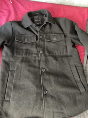Svart overshirt från Jack & Jones - Stilren svart overshirt från Jack & Jones med klassisk krage, två bröstfickor med lock och knappar samt sidofickor. Tillverkad i ett mjukt ull-liknande material och helfodrad för extra komfort. Perfekt lager-på-lager-plagg för en trendig look.