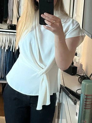 Vit knytblus med kort ärm - Stilren vit blus med knytband framtill och korta ärmar. Blusen har en mjuk, något glansig yta och en elegant drapering över bröstet. Perfekt till både jeans och kostymbyxor för en trendig look.