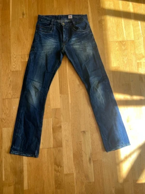 Blå raka jeans från Edwin - Säljer ett par blå jeans från Edwin med raka ben och slitna detaljer. Jeansen har klassisk femficksmodell, knappgylf och tydliga slitningar framtill för en cool, avslappnad look. Perfekta för dig som gillar streetwear och vintage vibe.