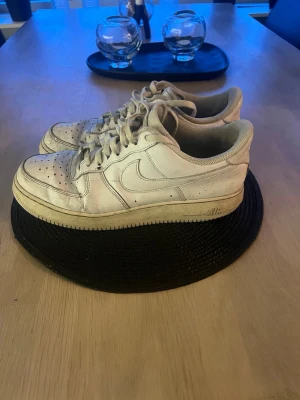 Nike Air Force 1 vita sneakers - Klassiska vita Nike Air Force 1 sneakers med lågt skaft och ikonisk swoosh på sidan. Skorna har perforerad tå för ventilation, tjock sula och är tillverkade i läder med textilfoder. Perfekta för en streetwear-look.