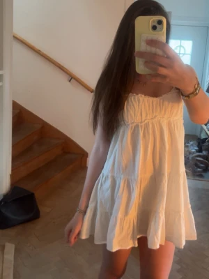 Vit kort sommarklänning med volang - Söt vit kortklänning med tunna axelband och volangdetaljer. Klänningen har en luftig passform och är perfekt för varma dagar. Tillverkad i ett lätt bomulls material som känns skönt mot huden. Enkel och stilren design som ger en fräsch look.