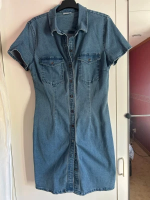 Jeansklänning från Noisy May - Snygg jeansklänning från Noisy May med kort ärm, knappar hela vägen framtill och två bröstfickor. Klassisk skjortkrage och figurnära passform💙