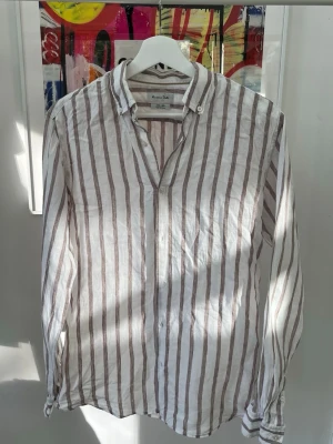 Randig linneskjorta från Massimo Dutti - Snygg vit och ljusbrun/guldig skjorta i 100% linne från Massimo Dutti med bruna vertikala ränder. Skjortan har klassisk krage, knäppning framtill och långa ärmar. Perfekt för en avslappnad och stilren look.