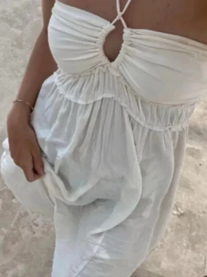 Vit långklänning med knyt från H&M - Säljer en vit långklänning med smala axelband som korsas framtill och en snygg cut-out detalj vid bysten. Klänningen har en luftig passform och är tillverkad i ett lätt tyg, perfekt för varma dagar.
