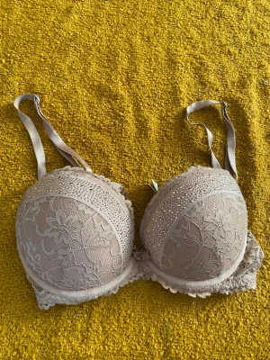 Beige push-up bh med spets och strass - Snygg beige push-up bh med spetsdetaljer och blommigt mönster. Kuporna är dekorerade med glittrande strass och har en liten rosett framtill. Axelbanden är justerbara och bh:n har hakknäppning bak.