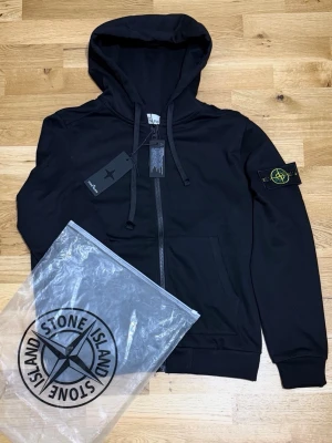 Stone Island Zip Hoodie - Säljer en svart hoodie från Stone Island med dragkedja framtill och klassisk logopatch på vänster ärm. Hoodien har huva med dragsko och ribbade muddar. Tillverkad i mjukt bomullsmaterial och levereras med originalpåse. TVEKA INTE ATT SKICKA EN BUD:)