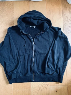 Svart zip-hoodie från Weekdy  - En klassisk svart hoodie med dragkedja framtill, huva med dragsko och två stora fickor på magen. Tröjan har ribbade muddar vid ärmslut och nederkant. Perfekt för en avslappnad och stilren look.