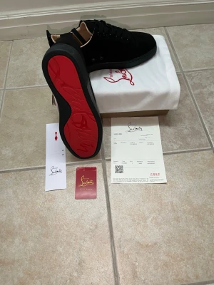 Svarta Christian Louboutin sneakers - Snygga svarta sneakers från Christian Louboutin med klassisk röd sula. Skorna har en låg siluett, svarta skosnören och ovandel i mocka. Perfekta för dig som vill sticka ut med exklusiva detaljer och ikonisk design.