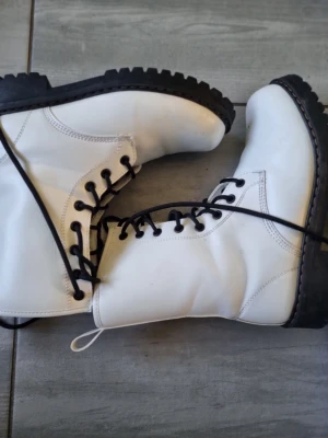 Vita snörade boots med grov sula - Säljer ett par vita boots med svart grov sula och svarta snören. Skorna har högt skaft och metallhål för snörningen. Perfekta för dig som vill sticka ut med en cool och edgy stil. Materialet är slätt och bootsen har en klassisk form.