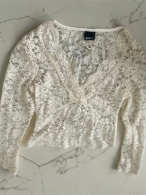 Vit spetsblus från Gina Tricot - Superfin vit blus i spets från Gina Tricot med v-ringning och långa ärmar. Blusen har ett romantiskt blommigt spetsmönster och är lätt croppad. Perfekt att styla med jeans eller kjol för en trendig look.