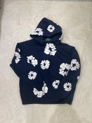 Blå hoodie med vita blommor Denim Tears - Snygg marinblå hoodie från Denim Tears med stora vita blommönster över hela plagget. Klassisk huva och känguruficka framtill. Tillverkad i mjukt bomullsmaterial som känns skönt mot huden. Perfekt för dig som vill sticka ut med en unik och trendig stil.