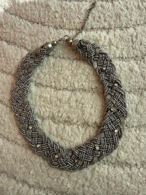 Flätat halsband med silverfärgade pärlor - Snyggt halsband med flätad design av små silverfärgade pärlor. Halsbandet har en metallkedja och justerbart lås. Pärlorna ger ett glittrigt och trendigt intryck, perfekt för att lyfta vilken outfit som helst. Materialet är metall och glas.