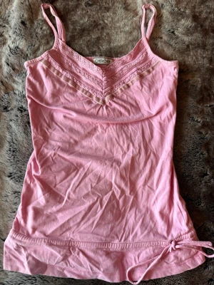 Rosa linnetopp från Cherokee - Söt rosa linnetopp från Cherokee med spetsdetaljer och smala axelband. Toppen har v-ringning och en dekorativ knytning i nederkanten. Perfekt för varma dagar och enkel att styla till jeans eller kjol.