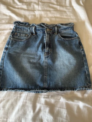 Blå jeanskjol med paperbag-midja - Snygg blå jeanskjol med paperbag-midja och rå kant nertill. Kjolen har klassiska fem fickor, bälteshällor och stängs med knapp och dragkedja framtill. Perfekt till vardags för en avslappnad men trendig look.